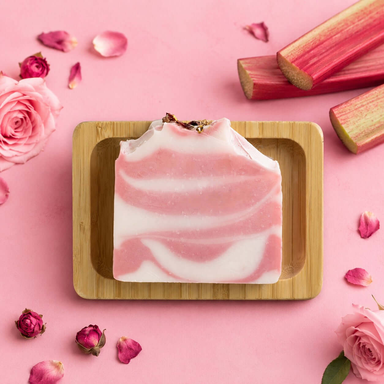 Bio Seife 'fruchtige Rose' auf Bambusholzablage, umgeben von Rosenblüten und Rhabarber.